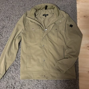 Grön jacka från J.Lindeberg - Snygg grön jacka från J.Lindeberg med dragkedja och knappar. Jackan har en stilren design med långa ärmar och en mjuk insida. Perfekt nu när de blir kallt igen eller till kommande vår!