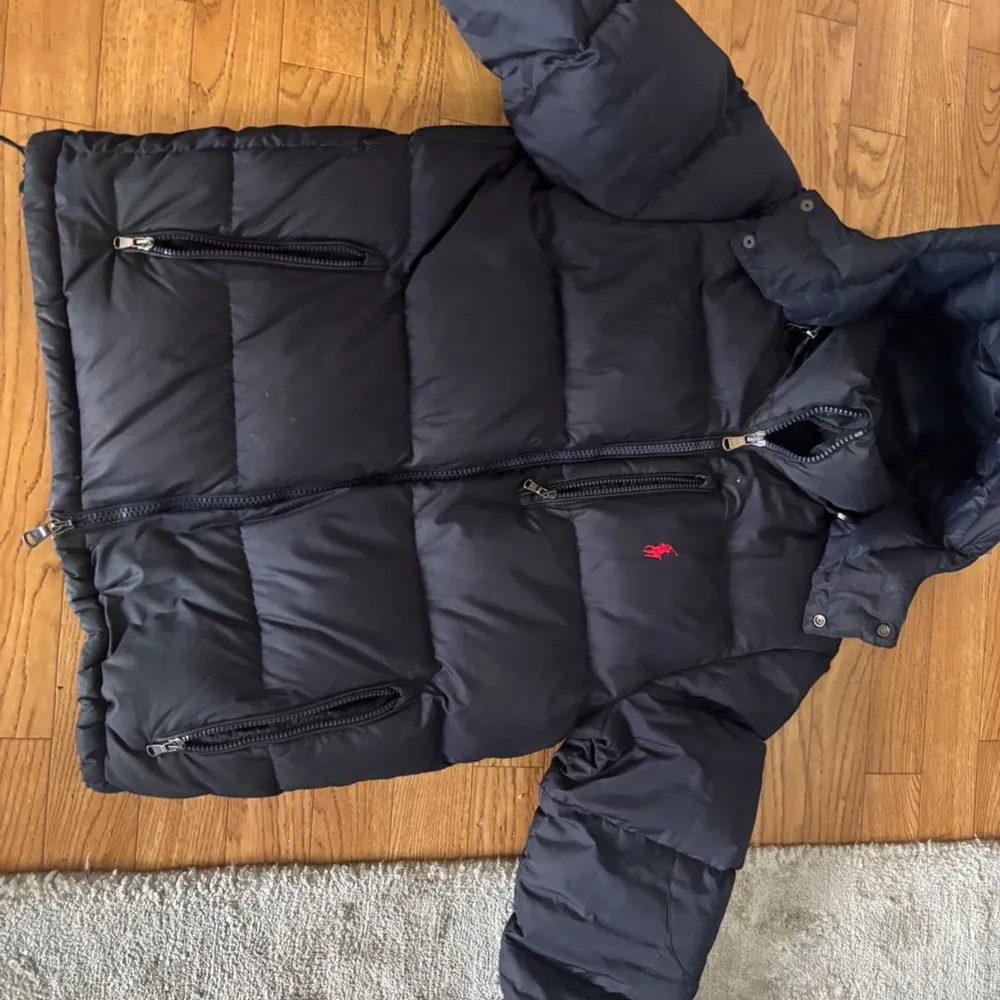 Säljer en svart dunjacka från Polo Ralph Lauren med dragkedja och flera praktiska fickor. Jackan har en broderad logga på bröstet och är perfekt för kyligare väder. Den är långärmad och har en klassisk design.. Takit.