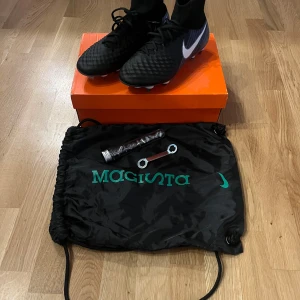 Svarta fotbollsskor från Nike - Säljer ett par svarta Nike fotbollsskor med snörning och hög ankelkrage för extra stöd. Skorna har en vit Nike-logga på sidan och är designade för optimal prestanda på planen. Perfekta för fotbollsspelare som söker både stil och funktion.