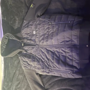 Svart cardigan från Stone Island - Säljer en svart cardigan från Stone Island med quiltad design och dragkedja. Jackan har en huva och en broderad logotyp på ärmen. Perfekt för en stilren look.