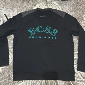 Svart tröja från Hugo Boss - Snygg svart tröja från Hugo Boss med turkos logga på bröstet. Tröjan har långa ärmar och en rund halsringning. Perfekt för en stilren look. XL men passar L