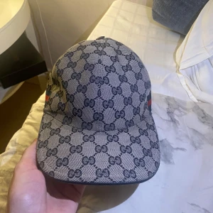 Mönstrad keps från Gucci - Snygg keps från Gucci med det ikoniska GG-mönstret i grått och svart. Kepsen har en klassisk design med böjd skärm och justerbar rem baktill. Perfekt för att ge din outfit en lyxig touch. Gick tyvärr inte att lägga till fler bilder så om du är intresserad och vill se mer så skickar jag.