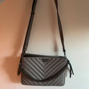 Grå axelväska med kedjedetalj - Snygg grå axelväska med justerbar rem och kedjedetalj framtill. Perfekt för att ge din outfit en stilren touch.