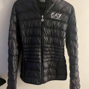 Svart pufferjacka från Emporio Armani - Snygg svart pufferjacka från Emporio Armani EA7 med dragkedja och huva. Det är storlek L men passar mig väldigt bra som vanlig viss har storlek S i jackor så skulle säga att den är väldigt liten i storleken. Andvänd få gånger.