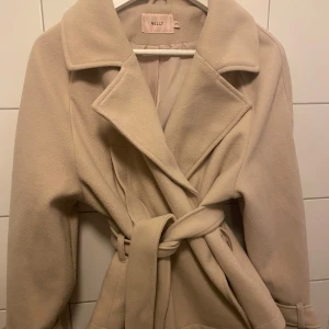 Beige kappa från Nelly - Snygg beige kappa från Nelly med knytbälte i midjan och breda slag. Den har långa ärmar med dekorativa knappar vid ärmsluten. Perfekt för kyligare dagar med sin stilrena design.