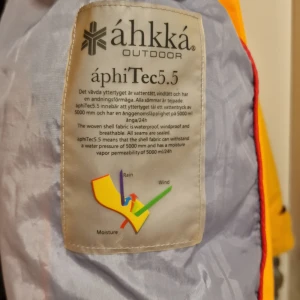 Gul vindjacka från Áhkká - Säljer en gul vindjacka och byxor från Áhkká med áphiTec5.5 teknologi som är både vattentät och vindtät. Jackan har en justerbar huva och dragkedja framtill. Perfekt för utomhusaktiviteter i alla väder.