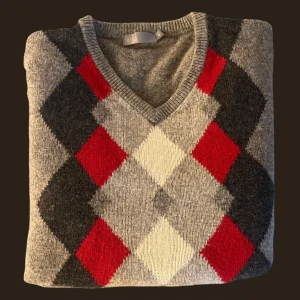 Grå tröja med argylemönster - Säljer en stilren grå tröja med klassiskt argylemönster i rött, svart och vitt. Tröjan har en v-ringad hals och långa ärmar, perfekt för en elegant look. Passar bra till både vardag och mer uppklädda tillfällen.
