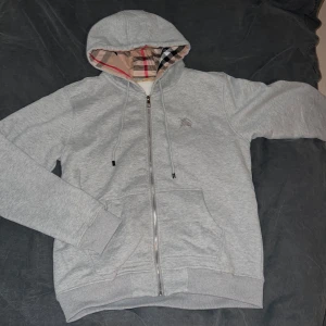 Grå hoodie från Burberry - Säljer en stilren grå hoodie från Burberry med dragkedja och klassiskt rutigt mönster i huvan. Tröjan har en broderad logga på bröstet och ribbade muddar. Perfekt för en avslappnad och trendig look. Fits big S and M and small L