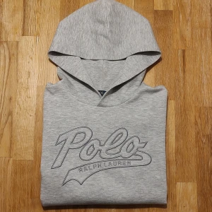 Ralph Lauren grå hoodie Polo - Supersnygg hoodie från Ralph Lauren, gråmelerad med broderad logga över bröstet. Stl 14-16år, sitter ungefär som en vuxen XS eller XXS, se mått! Riktigt fint skick. Lite "glatt" känsla på tyget. Mått: Armhåla till armhåla 50cm, bredd längst ner 44cm, längd från mitt på axeln 58cm.