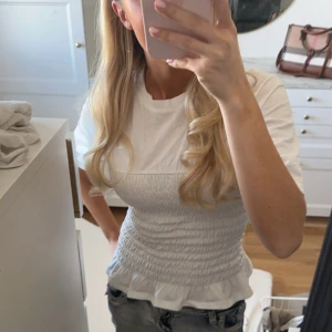Vit smockad topp - Säljer en söt vit topp med smockad design och korta ärmar. Toppen har en volangdetalj vid nederkanten som ger en feminin touch. Perfekt för en stilren look.