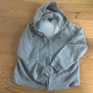 Grå hoodie från Polo Ralph Lauren - Säljer en grå hoodie från Polo Ralph Lauren med dragkedja och huva. Den har ett litet broderat logotypmärke på bröstet och är fodrad med mjukt material för extra komfort. Perfekt för kyliga dagar!