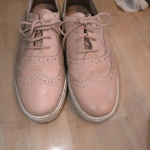 Rosa brogue-inspirerade skor - Säljer ett par rosa brogue-inspirerade skor med snörning och dekorativa perforeringar. Skorna har en vit sula med räfflad design för extra grepp. Perfekta för en stilren look.
