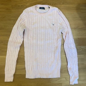 Rosa kabelstickad tröja från GANT - Säljer en stilren rosa stickad tröja från GANT i stretchig bomull. Tröjan har ett klassiskt kabelstickat mönster och långa ärmar. Perfekt för en avslappnad och snygg look. 
