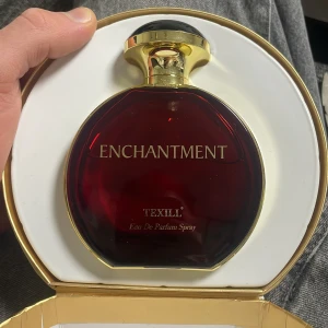 Enchantment Pour Homme Eau De Parfum från  - Elegant parfymflaska i djupröd färg med gyllene detaljer och svart topp. Flaskan har en rund form och kommer i en matchande röd förpackning med ett stilfullt vingmotiv. Perfekt för den som söker en sofistikerad doftupplevelse. Ca 90ml kvar!