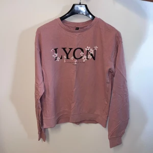 Rosa tröja från H&M Divided - Säljer en söt rosa tröja från H&M Divided med texten 'LYON' och blommiga detaljer. Tröjan har långa ärmar och en avslappnad passform. Perfekt för en casual look!