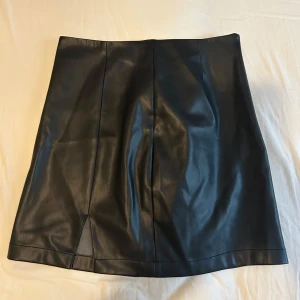 Svart skinnkjol från BikBok - Snygg svart skinnkjol från BikBok i en klassisk kort modell. Kjolen har en stilren design utan onödiga detaljer. Har en liten slits. 