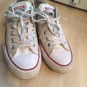 Vita Converse All Star sneakers - Säljer ett par klassiska vita Converse All Star sneakers med snörning och vit tåhätta. Skorna har en röd rand längs sulan och är perfekta för en avslappnad stil.