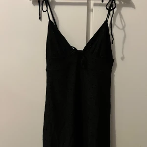 Svart sommarklänning - Snygg svart klänning med smala axelband och knytband framtill. Klänningen har ett diskret mönster. 