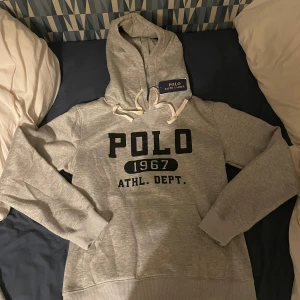 Ralph Lauren hoodie - Nu har du chansen att äga en ny grå hoodie från POLO RALPH LAUREN. Den kombinerar stil och komfort med klassisk design, framficka och dragsko i huvan. Prydd med trycket "POLO 1967 ATHL. DEPT." i svarta bokstäver på framsidan. Perfekt för både vardagsbruk och aktiva dagar. Missa inte chansen att lägga vantarna på detta högkvalitativa plagg! Helt ny ! Priset kan diskuteras vid en snabb och smidig affär. 
