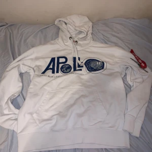 Vit hoodie från Alpha Industries - Säljer en vit hoodie från Alpha Industries med blått Apollo-tryck på framsidan. Hoodien har en klassisk känguruficka och en röd 'Remove Before Flight'-tag på ärmen. Perfekt för en avslappnad stil.