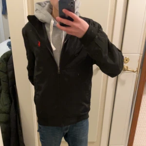 Svart jacka från Ralph Lauren - Säljer en stilren svart jacka från Ralph Lauren med dragkedja och röd broderad logga på bröstet. Jackan är i storlek M/S.skriv vid funderingar.