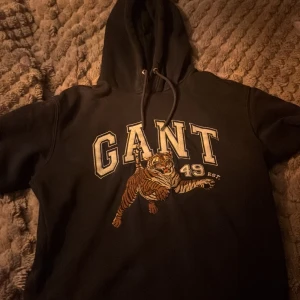 Svart hoodie från GANT med tigertryck - Snygg svart hoodie från GANT med ett coolt tigertryck och texten 'GANT 49 EST.' på framsidan. Perfekt för en avslappnad stil med en touch av attityd. Den har en klassisk huva med dragsko.