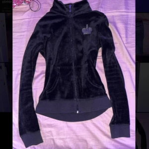 Svart vintage velourjacka från Juicy Couture - Snygg svart velourjacka från Juicy Couture med dragkedja och glittrande detaljer. Jackan har en tight passform och långa ärmar. Perfekt för en trendig look med en touch av glamour.