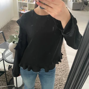 Svart blus med volangdetaljer - Snygg svart blus med volangdetaljer längs ärmarna och spetsinslag. Blusen har långa ärmar och knappar i ryggen. Perfekt för en stilren look.