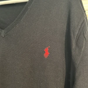 Svart tröja från Polo Ralph Lauren - Säljer en stilren svart tröja från Polo Ralph Lauren i 100% Pima bomull. Tröjan har en klassisk v-ringning och det ikoniska röda Polo-logotypen broderad på bröstet. Perfekt för en elegant och avslappnad look.