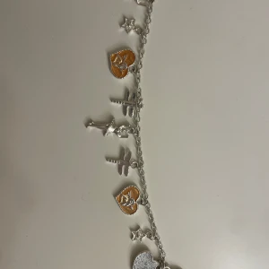 Silverfärgat armband med berlocker - Charmigt silverfärgat armband med olika berlocker, inklusive stjärnor, hjärtan och kors. Några av hjärtberlockerna har en orange detalj som ger en fin kontrast. Perfekt för att ge en personlig touch till din stil.