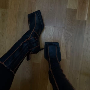 Miista svarta boots - Snygga svarta boots med fyrkantig tå och kontrasterande sömmar i orange röd. De har en unik design med spetsig klack och dragkedja på sidan för enkel på- och avtagning. Perfekta för att ge en edgy touch till din outfit. Lite slitna på skinnet på klacken. Ord 450 euro