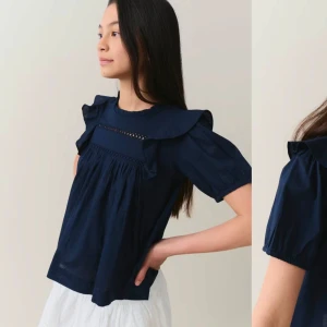 Mörkblå blus med volangdetaljer - Charmig mörkblå blus med volangdetaljer vid axlarna och halsen. Blusen har korta ärmar och en luftig passform, perfekt för en stilren look. Den har även en fin struktur vid bröstet som ger extra stilpoäng.