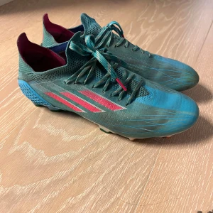 Blåa fotbollsskor från Adidas - Snygga blåa fotbollsskor från Adidas med röda detaljer och snörning. Skorna har en strömlinjeformad design med en robust sula för bra grepp på planen. Perfekta för fotbollsträning och matcher.