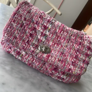 Rosa rutig axelväska med kedja - Säljer en snygg rosa rutig axelväska med en stilren kedja. Väskan har ett vridlås i metall och är perfekt för att ge din outfit en färgklick. Insidan är ljusblå med en liten logga. Perfekt för att bära dina nödvändigheter med stil.