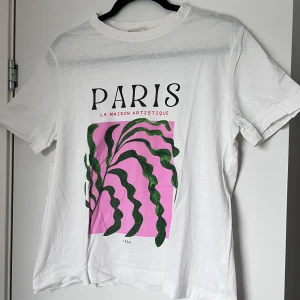 Vit t-shirt med tryck 'Paris' - Säljer en vit t-shirt med ett färgglatt tryck av texten 'Paris' och en konstnärlig design i grönt och rosa. Perfekt för en avslappnad stil med korta ärmar och rund halsringning. Storlek: S. Axel: 41cm. Längd: 61cm. 🩷💚