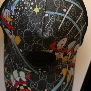 Gucci x Disney keps med kalle anka motiv - Snygg keps från Gucci med ett färgglatt Disney-motiv. Kepsen har ett svart märke framtill och ett unikt mönster med stjärnor och karaktärer. Perfekt för den som vill sticka ut med stil.