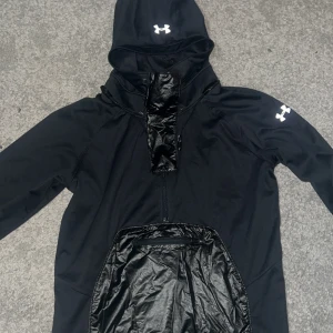 Svart vindjacka från Under Armour - Säljer en svart vindjacka från Under Armour med huva och dragkedja. Jackan har en stor ficka framtill och är långärmad. Perfekt för sportiga aktiviteter och håller dig skyddad mot vinden.