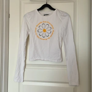 Vit långärmad topp från BikBok - Säljer en söt vit långärmad topp från BikBok med ett tryck av en stor blomma och text i gult och svart.🌼Helt ny köpte för två somrar sen i storlek XS.