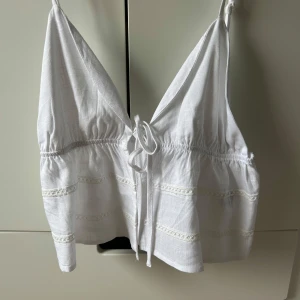 Vit linne topp med spetsdetaljer - Säljer en söt vit linne topp med smala axelband och spetsdetaljer. Toppen har en knytning framtill och en luftig passform, perfekt för varma dagar.