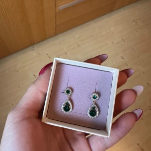 Gröna örhängen från Lily and Rose - Eleganta gröna örhängen och matchande halsband från Lily and Rose. Båda smyckena har en droppformad grön sten omgiven av gnistrande stenar i en guldfärgad infattning. Perfekt för student, bröllop, midsommar och balen. Säljer de separata.