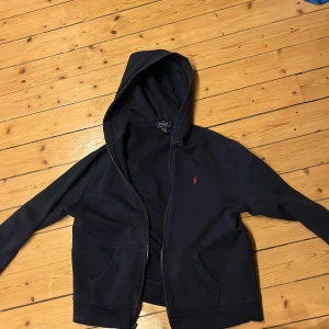 Mörkblå hoodie från Ralph Lauren - Säljer en snygg mörkblå hoodie från Ralph Lauren med dragkedja och huva. Den har en röd broderad logga på bröstet och praktiska fickor framtill. Perfekt för en avslappnad stil. Nypris ca 1200kr passar dig som är 160-170