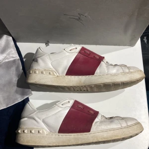 Valentino Garavani sneakers med röd detalj - Snygga Valentino Garavani sneakers i vitt med en bred röd rem över snörningen. Skorna har en unik design med nitar runt sulan och är perfekta för en stilren look. I rätt dåligt skick men fortfarande användbara, sulan har reparerats en gång men det borde inte vara något problem. Såklart äkta, finns äkthetsgaranti!