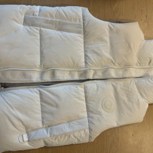 Vit dunväst från Canada Goose - Säljer en stilren vit dunväst från Canada Goose. Västen har en dragkedja framtill och är perfekt för kyligare dagar. högkvalitativa material. 1.1k