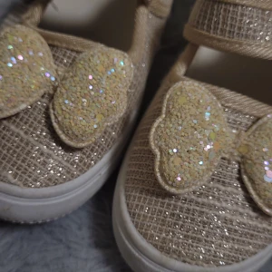 Beige glittriga barnskor - Söta beige barnskor med glittriga detaljer och kardborreband för enkel på- och avtagning. Skorna har en vit sula och dekorativa glitterdetaljer på ovansidan som ger en lekfull touch. I storlek 27. De är I helt nytt skick, använda en gång till en fest annars helt nya, har inte kommit till mer användning än så. Pris kan rekommenderas!