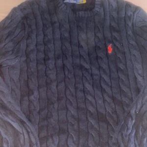 Mörkblå stickad tröja från Ralph Lauren - Säljer en snygg mörkblå stickad tröja från Ralph Lauren med det klassiska röda logotypen på bröstet. Tröjan har ett kabelstickat mönster och långa ärmar. Perfekt för kyliga dagar!