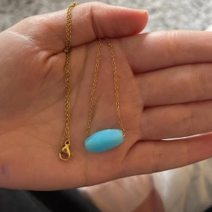 Halsband med blå sten - Elegant halsband med en ljusblå oval sten hängande från en tunn guldfärgad kedja. Perfekt för att lägga till en färgklick till din outfit.