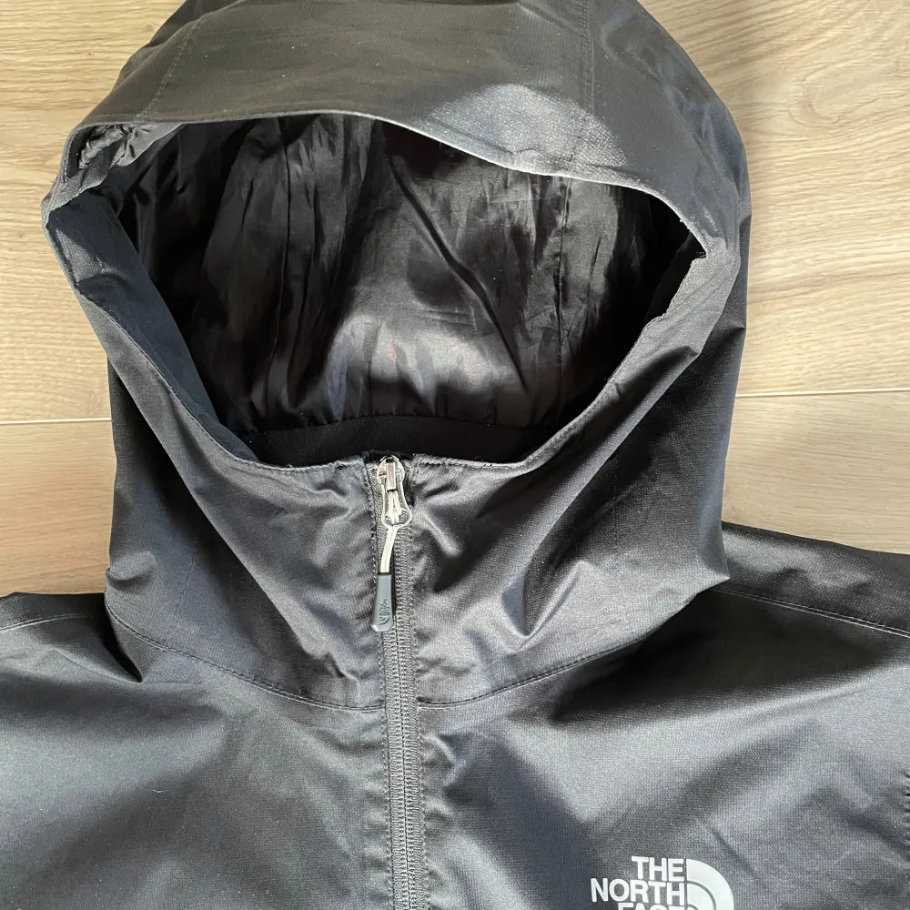 Säljer en svart regnjacka från The North Face med huva och dragkedja. Jackan har en stilren design och är perfekt för regniga dagar. Den är tillverkad i ett slitstarkt material och har The North Face-loggan på bröstet och ryggen.. Takit.