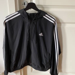 Svart vindjacka från Adidas -  Svart vindjacka från Adidas som aldrig använts. Jackan har dragkedja och huva, perfekt för en sportig look. Den är tillverkad i lätt syntetmaterial, 100 % polyester. Sidfickor.  Storlek S, 34-36. 