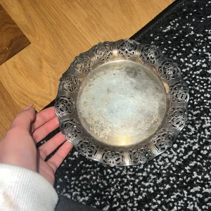 Dekorativ metalltallrik med utskuret mönster - Säljer en dekorativ metalltallrik med ett vackert utskuret mönster längs kanten. Tallriken har en antik finish och är perfekt som prydnad eller serveringsfat. Den har en rund form och en elegant design som passar i de flesta hem. Perfekt för dig som gillar vintage-stil!