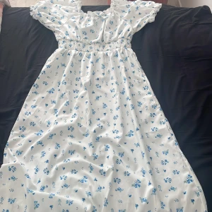 Vit blommig klänning från Shein - Charmig vit klänning från Shein med blått blommönster. Klänningen har korta ärmar med  volangeroch en söt rosett vid halsen. Perfekt för somriga dagar med sin lätta och luftiga design.